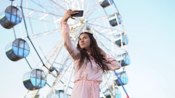 a Girl with Long Hair in a Dress Makes Selfie Using a Smartphone Standing Near the Ferris Wheel