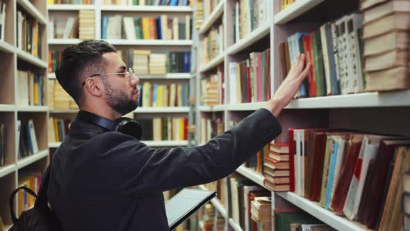 Student Choosing Books in Library According to List on Tablet alt