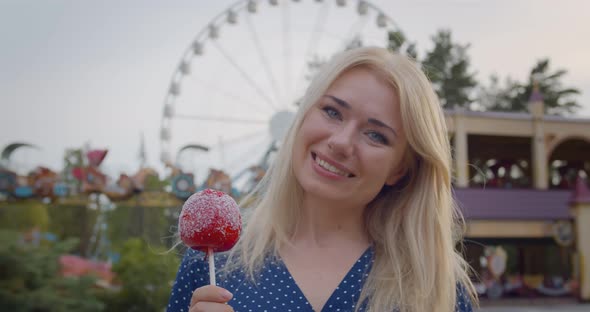 Portrait of Beautiful Young Woman in Amusement Park Holding Red Caramel Apple Looking at Camera