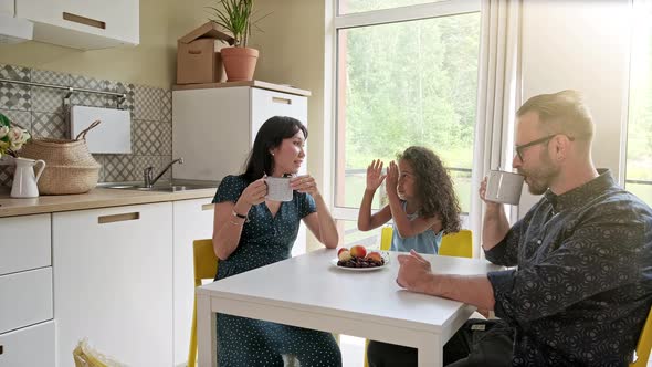 Beautiful Family with Kid Sitting at the Table Drinking Coffee at New Home Around Cardboard Boxes