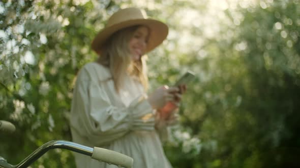 Portrait of Blonde Woman in Hat Enjoy Success on Mobile Phone of White Apple Blossoms