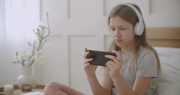 Teen Girl Is Listening Music By Headphones Connected with Smartphone By Wifi, Sitting at Her Room at