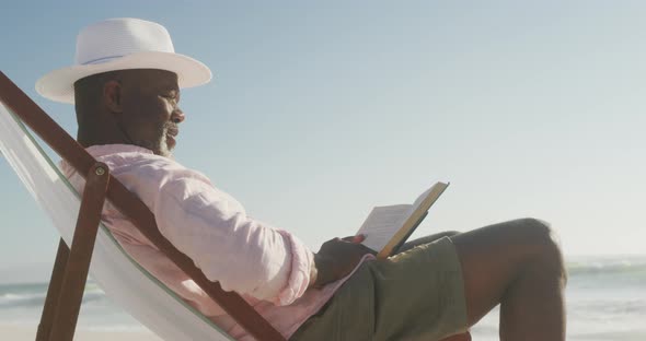 Smiling senior african american man lying on sunbed and reading book on sunny beach