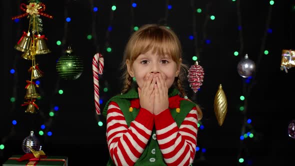 Kid Girl in Christmas Elf Santa Claus Helper Costume Looks at Camera Makes Big Eyes Covering Mouth