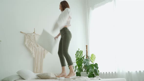 Young Funny Woman Jumps on the Bed with a Pillow in the Bedroom