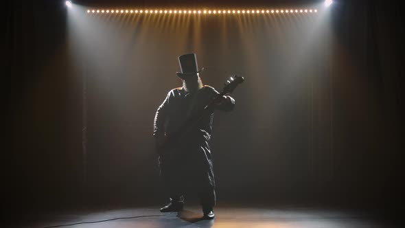 Silhouette of a Man with a Beard and a Hat Who Plays the Electric Guitar on Stage in a Dark Studio