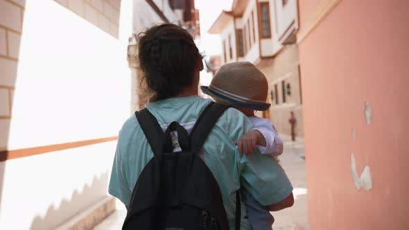 Mother with Backpack Carries Little Son Walking on Street