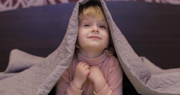 Little Cheerful Girl Hides Under Blanket and Watching TV. Concept of Kids Sleep