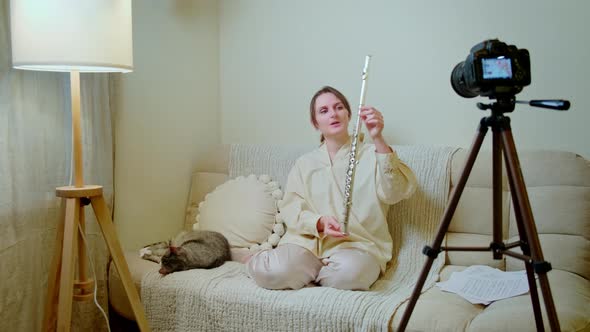 Happy woman musician with flute looking at video camera at home on sofa in living room