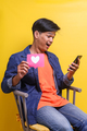 Young surprised Asian man sitting on the chair holding love sign for social media - PhotoDune Item for Sale