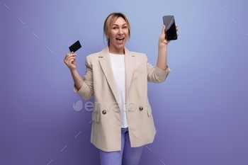 close-up of a joyful blonde woman in a jacket with a smartphone and a money card on a studio