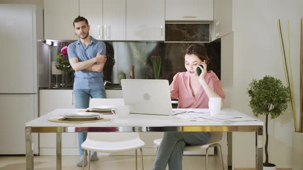 A Busy Woman is Working and Talking on the Phone While Her Husband is Standing and Looking at Her