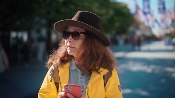 A Mature Adult Woman Uses the Phone and Looks Around While Walking at the City