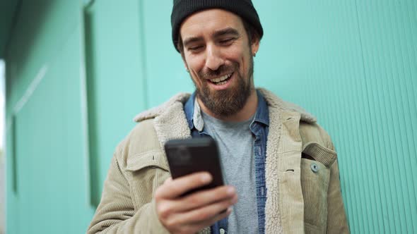 Positive bearded man wearing hat typing by phone and talking