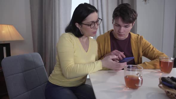 Teen Brunette Caucasian Boy Teaching Adult Mother in Eyeglasses To Use Smartphone. Patient Son