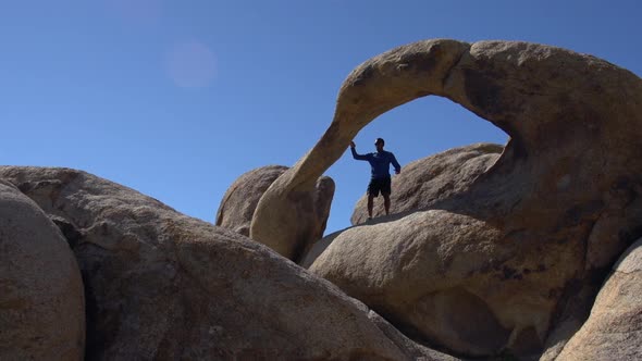 A young man trail running with his dog past a natural rock arch in a mountainous desert.