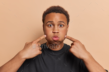gers at cheeks holds breath foolishes around dressed in casual black t shirt isolated over brown background. Human facial expressions concept