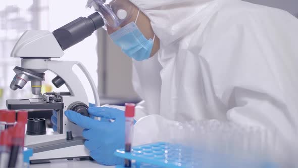 Scientist in PPE suite doing some research and looking through a microscope in laboratory