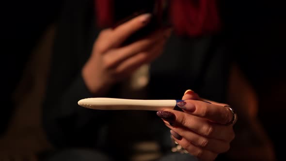 Young Woman Sits in Room in the Evening Holding Pregnancy Test and Taking Photo of It By Mobile