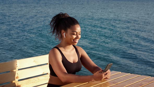 Young Positive African American Woman in Wireless Headphones Web Surfing on Smartphone Resting at