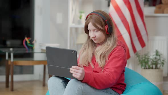 Confident Asian Teenage Girl in Headphones Using Tablet at Home with Blurred American Flag at