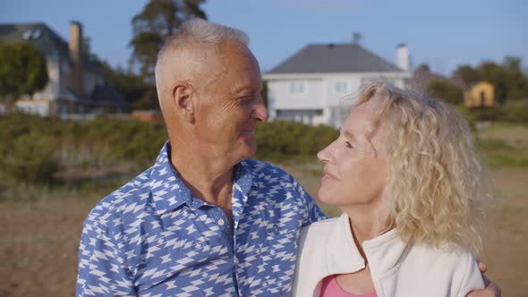 Happy Senior Couple Hugging on Sandy Beach Over Country House Background