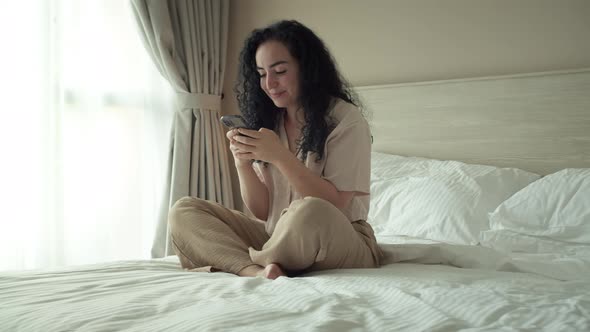 Cheerful Young Indian Woman Using Smartphone While Sitting on Bedroom