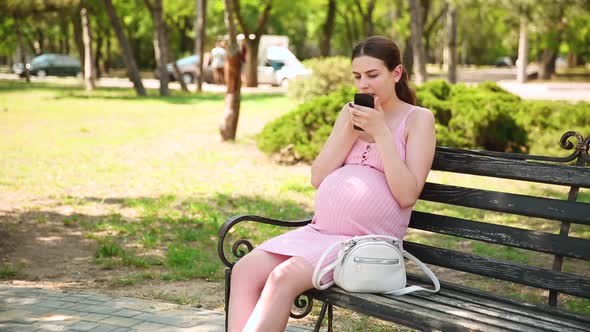 Pregnant Woman Paints Her Lips in the Summer Sunny Day in a City Park on a Bench