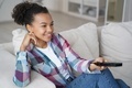 Smiling mixed race teen girl watching television series, changes TV channel, sitting on sofa at home - PhotoDune Item for Sale
