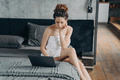 Focused woman wrapped in towel after shower works on laptop sitting on bed in morning. Distant work - PhotoDune Item for Sale