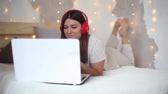 A Beautiful Sexy Girl in Pajamas Listening to Music on Headphones in Red Using a Laptop of White