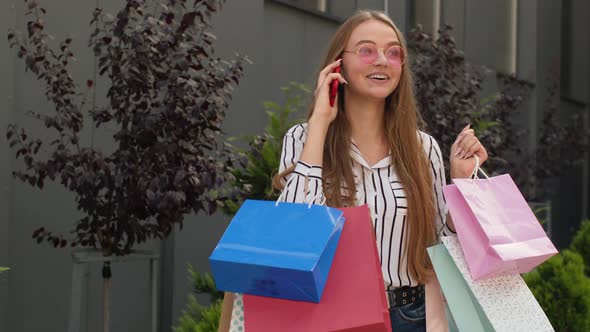 Positive Young Girl with Bags Talking on Mobile Phone About Sales in Shopping Mall in Black Friday