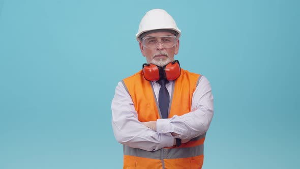 Senior man engineer with a helmet and work vest puts on protective glasses and folds arms on chest