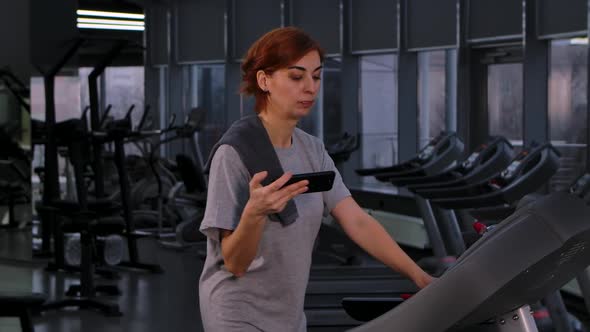 Young Woman Doing Speedwalk on Treadmill Looks at Smartphone Then Shocked and Excited