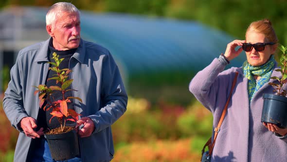 Portrait Of Retiree Couple With Potted Plants Walking Through Garden Spring Park. Selective Focus Sh