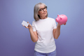 a woman of mature years with gray hair holds money capital from a piggy bank on a studio background - PhotoDune Item for Sale