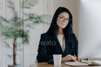 Serious female office worker in formal attire, wearing glasses, busy with online work, at her desk,