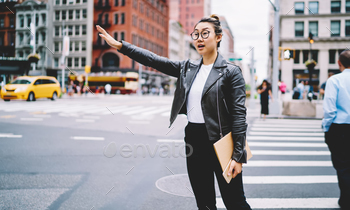 Trendy dressed hipster girl in optical spectacles waving at street with traffic