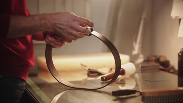 A Man Ties Up the Brand New Leather Belt By the Table in a Workshop