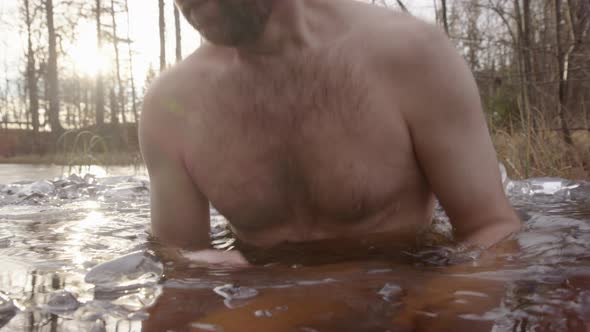 A handsome bearded man sits down into a bathing hole in a frozen lake
