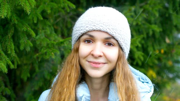 Blond Girl in Knitted Hat Smiles By Fur Tree in Falling Snow