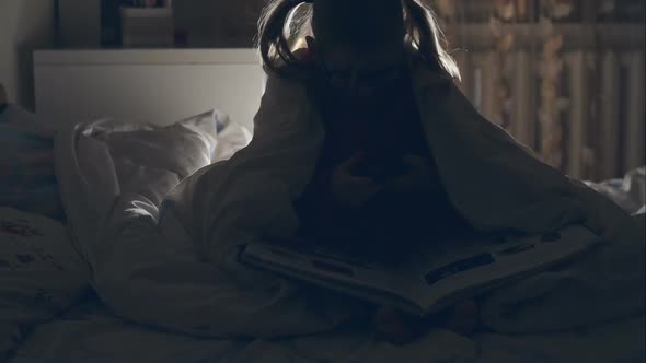 A Young Girl Reading a Book Under the Covers with a Flashlight. RAW Video Record.