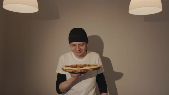 A Young Chef in a Bandana Holds a Pizza in a Cafe and Sniffs It Enjoying the Aroma