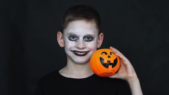 portrait of a cheerful caucasian boy 8-9 years old with zombie makeup on his face holding a pumpkin 