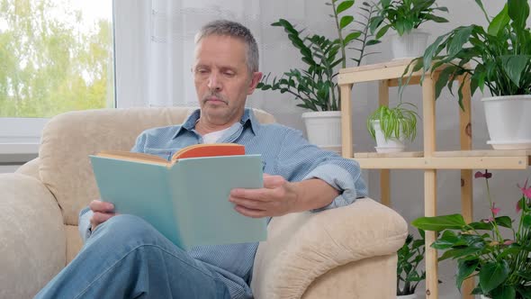 Senior Man Reads a Book While Sitting on Armchair at Home