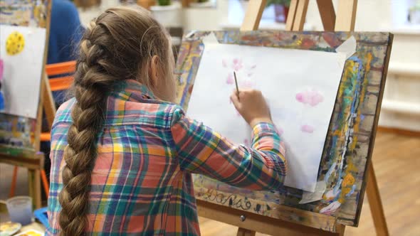 Young Cute Female Artist Is in an Art Studio, Sitting Behind an Easel and Painting on Canvas