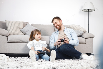 syndrome at home together. Man and girl sitting on a floor near sofa and holding joysticks. Bearded man wearing blue shirt and girl white shirt.