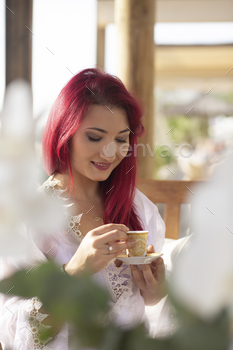 girl with red hair gown on a summer terrace among plants laughs drinks coffee,resting,morning coffee