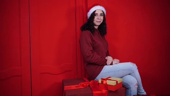 Young Attractive Woman in Santa Claus Hat Sits on Gifts Near Red Wall