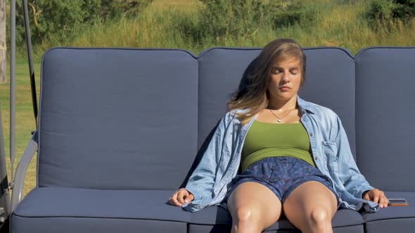 A young woman in shorts relaxes outside on a patio swing with her eyes closed. A gentle sea breeze b